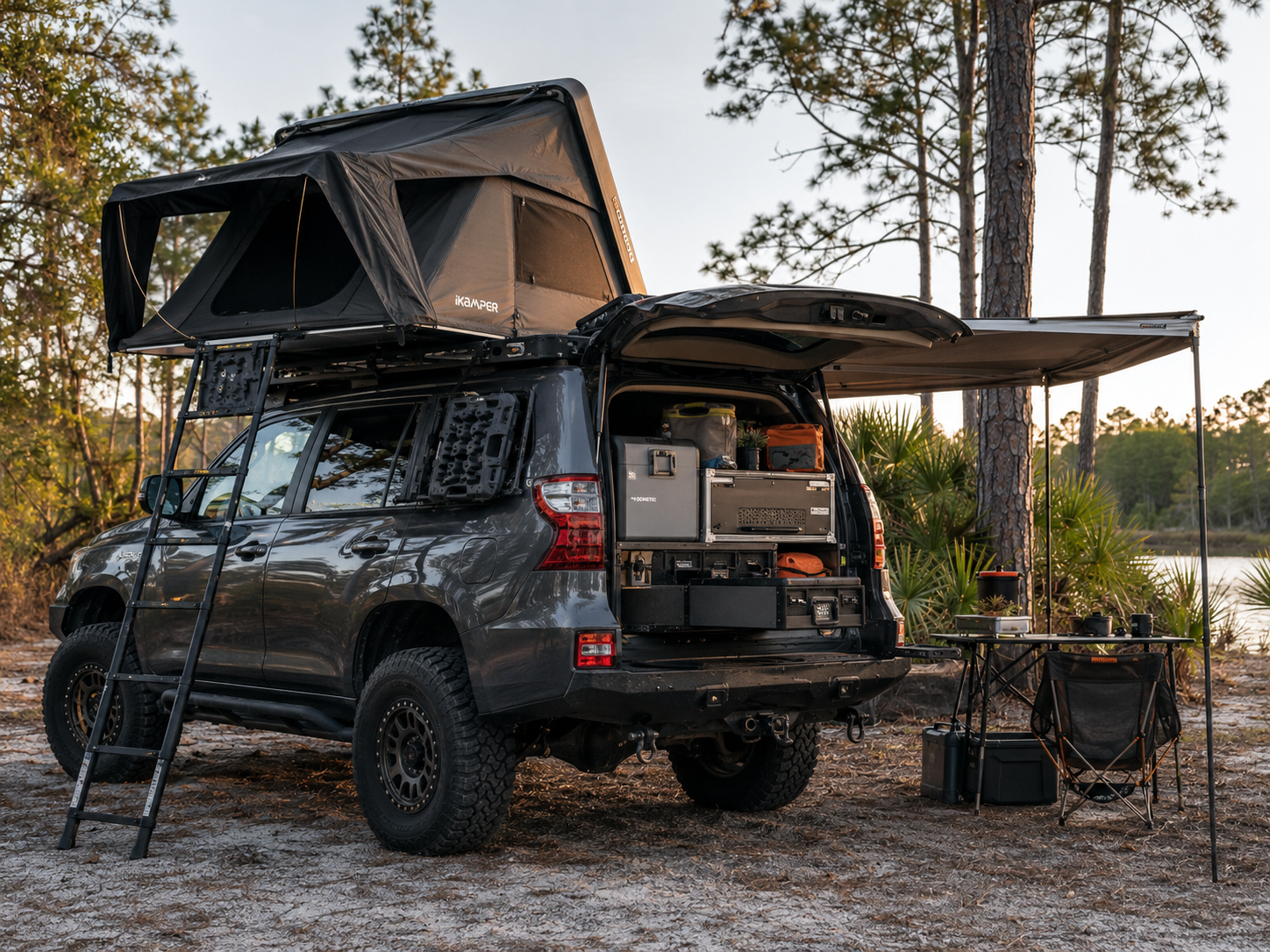 Lifted gray Lexus GX overland project with iKamper rooftop tent and awning deployed at a Florida pine forest lakeside campsite at golden hour
