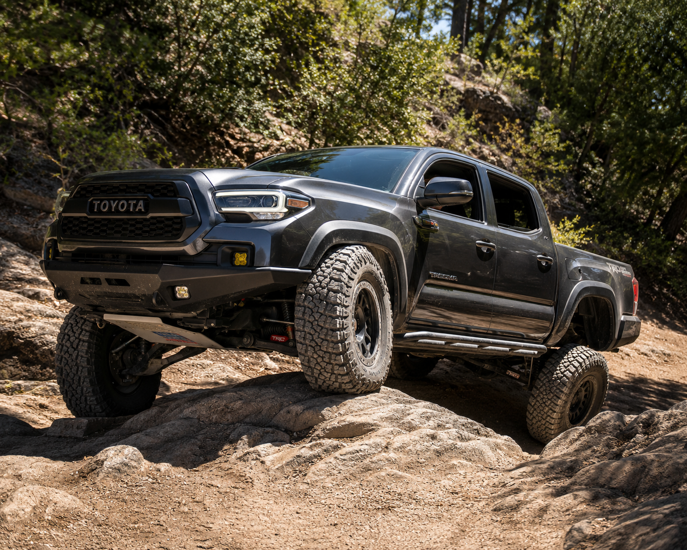 Lifted Toyota Tacoma with off-road suspension crawling over rocks on a wooded trail