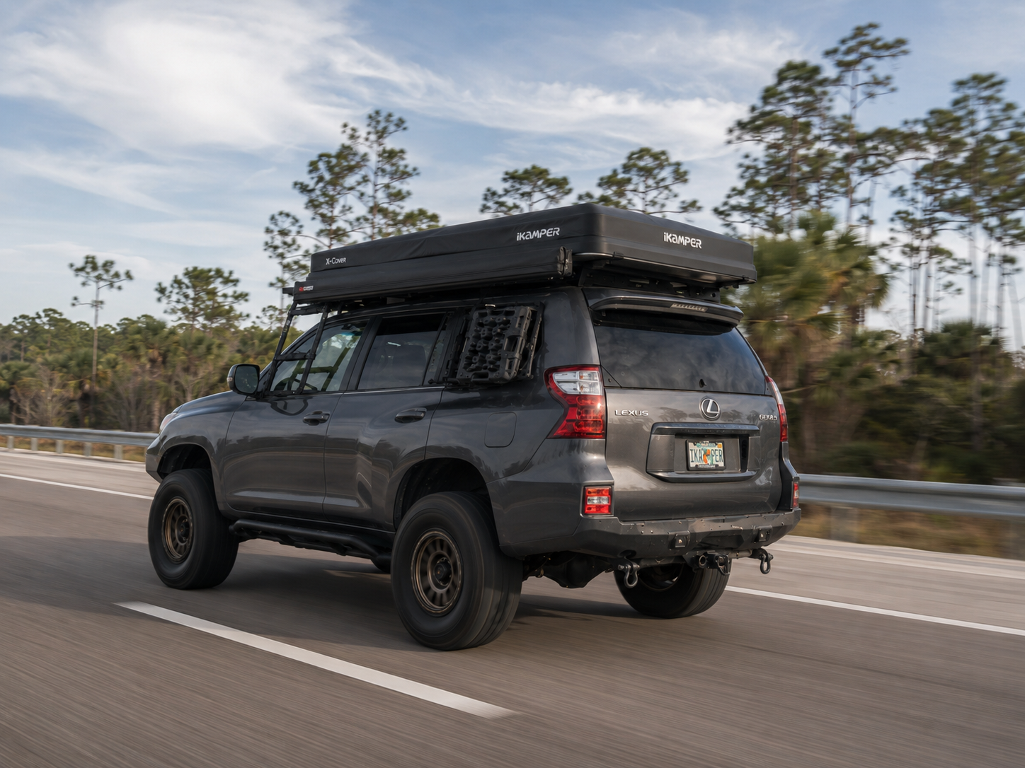 Lifted gray Lexus GX overland project with iKamper X-Cover rooftop tent driving on a Florida highway lined with pine trees